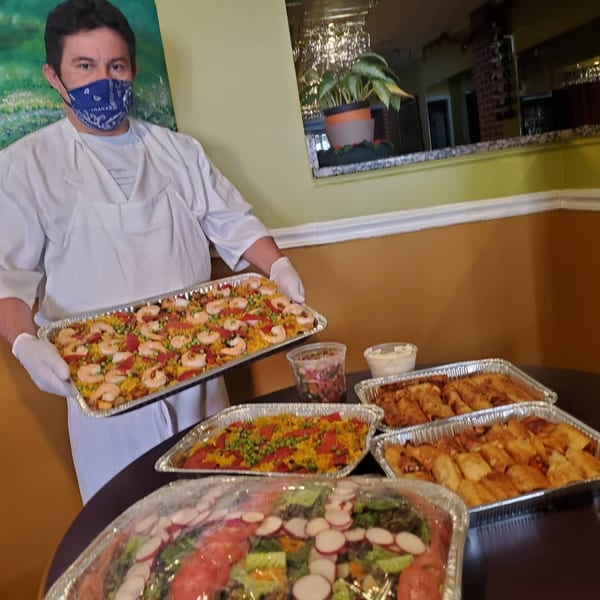Chef in a mask and white uniform holding a large tray of paella, with other Spanish dishes on a table.