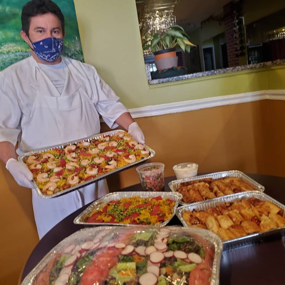 Chef in a mask and white uniform holding a large tray of paella, with other Spanish dishes on a table.