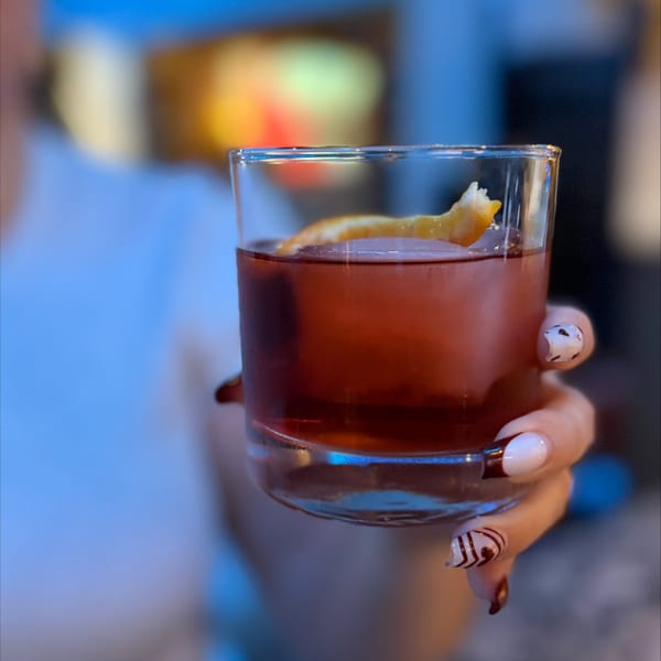 A person holds a short glass with a red cocktail and an orange peel garnish.