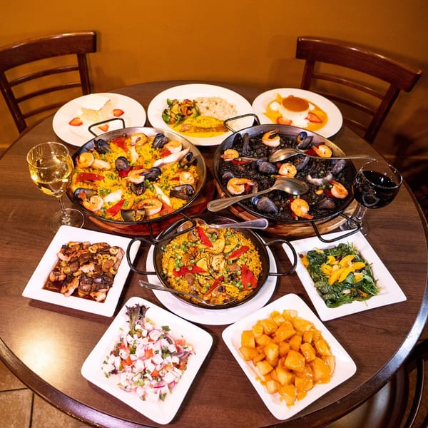 A table laden with various Spanish dishes, including paellas, tapas, and desserts, with wine glasses.