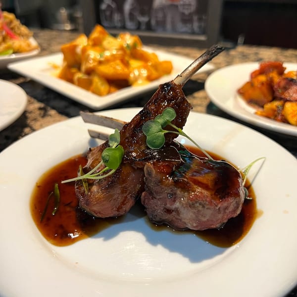 Two lamb chops with a dark glaze and microgreens on a white plate, with other dishes blurred in the background.
